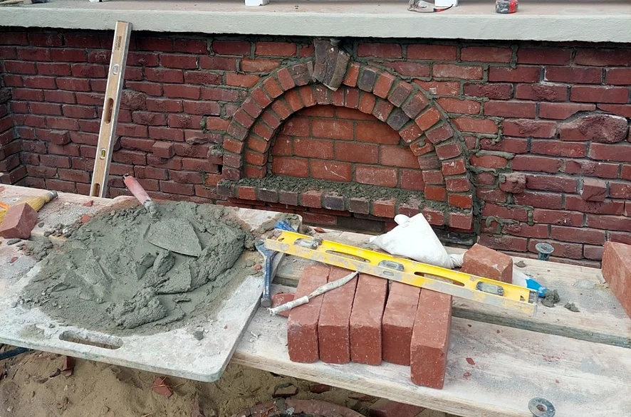 Trowel in fresh mortar on a mud board next to new brick and a level, in front of an arched brick repair under a porch